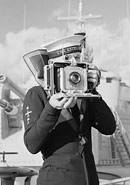 A photograph of a naval photographer attached to HMS Excellent poses with his Speed Graphic camera on the deck of a warship in port.